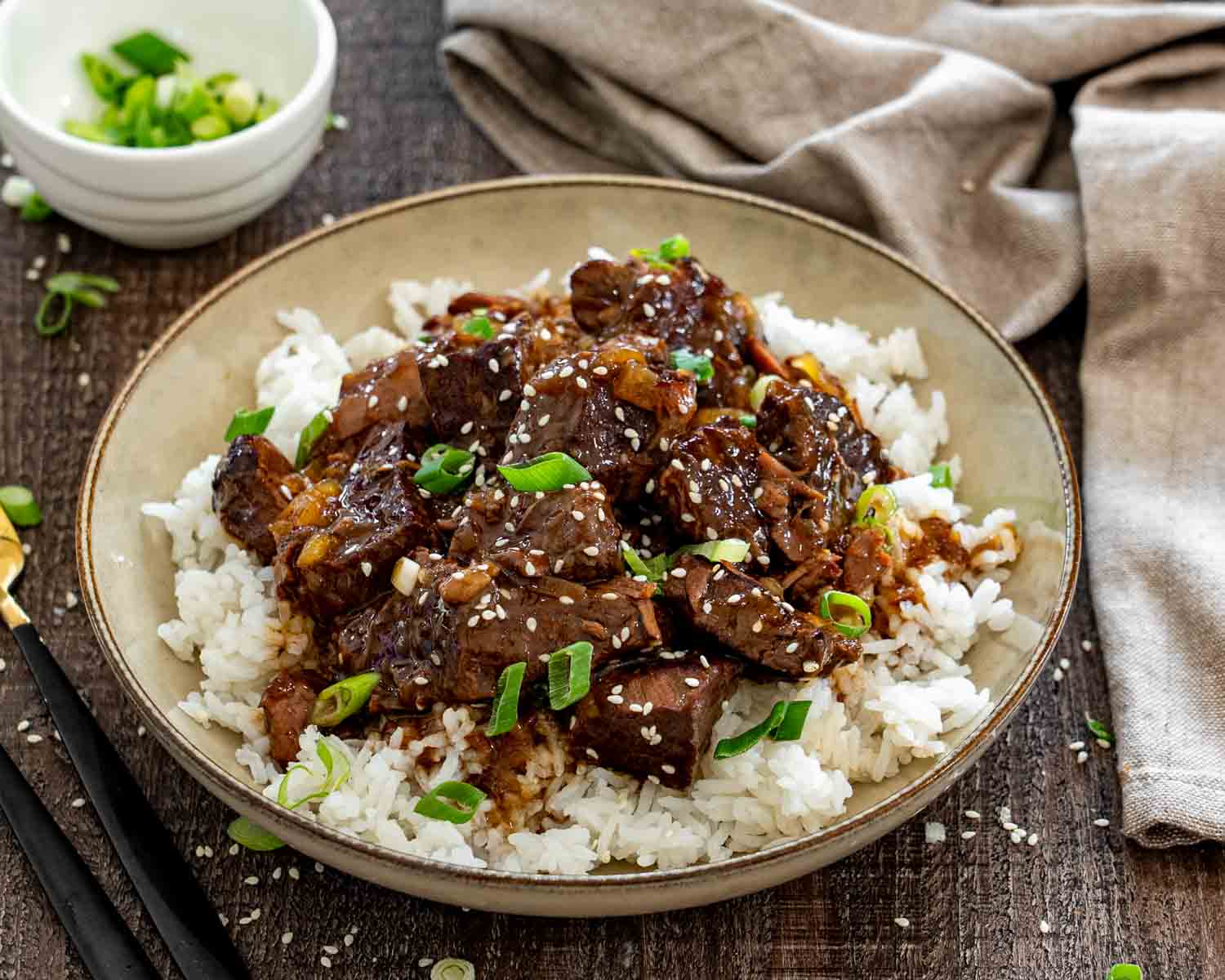 Asian braised beef piled over steamed white rice, garnished with sesame seeds and green onions in a shallow bowl. (