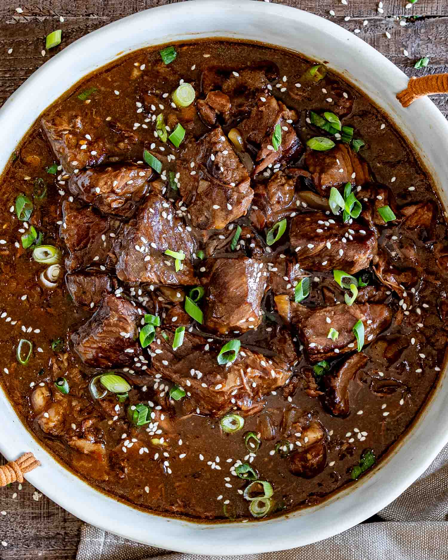 Overhead close-up of tender braised beef in rich brown sauce, finished with sesame seeds and sliced green onions.