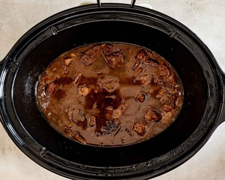 Overhead view of tender beef and onions simmering in a rich brown sauce with star anise inside a slow cooker.