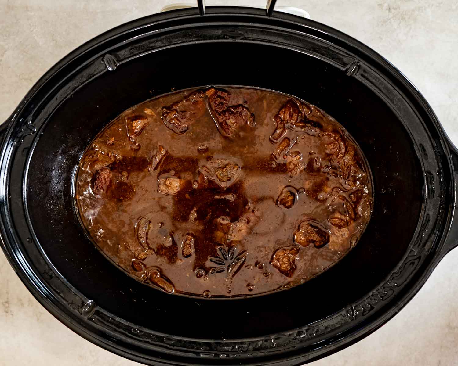 Overhead view of tender beef and onions simmering in a rich brown sauce with star anise inside a slow cooker.