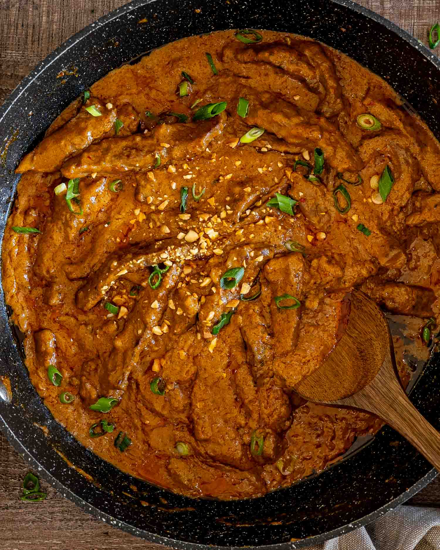 Overhead view of curried beef strips simmering in creamy peanut sauce, garnished with chopped peanuts and green onions.