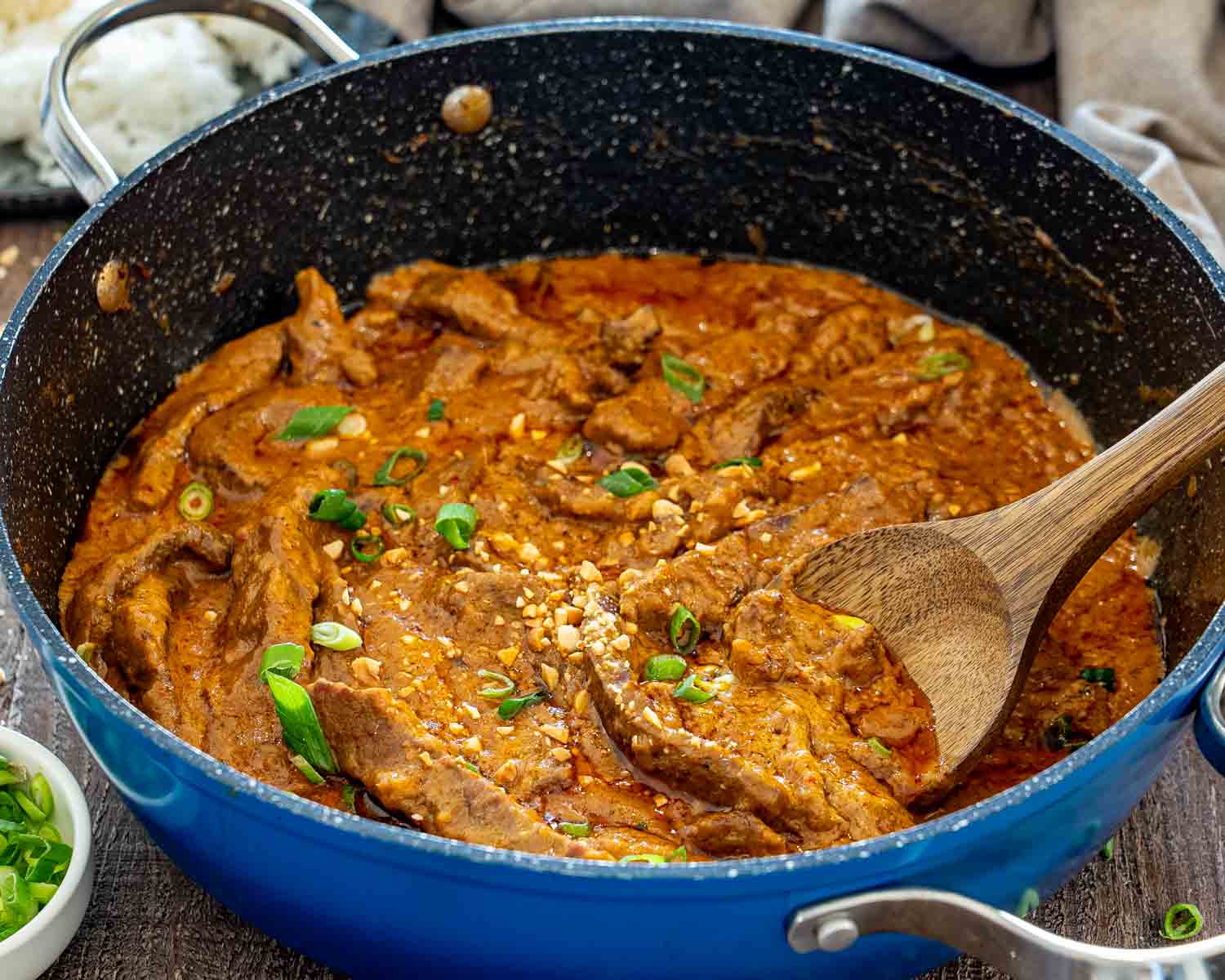 Close-up of curried beef simmering in creamy peanut sauce inside a blue skillet with chopped peanut garnish.