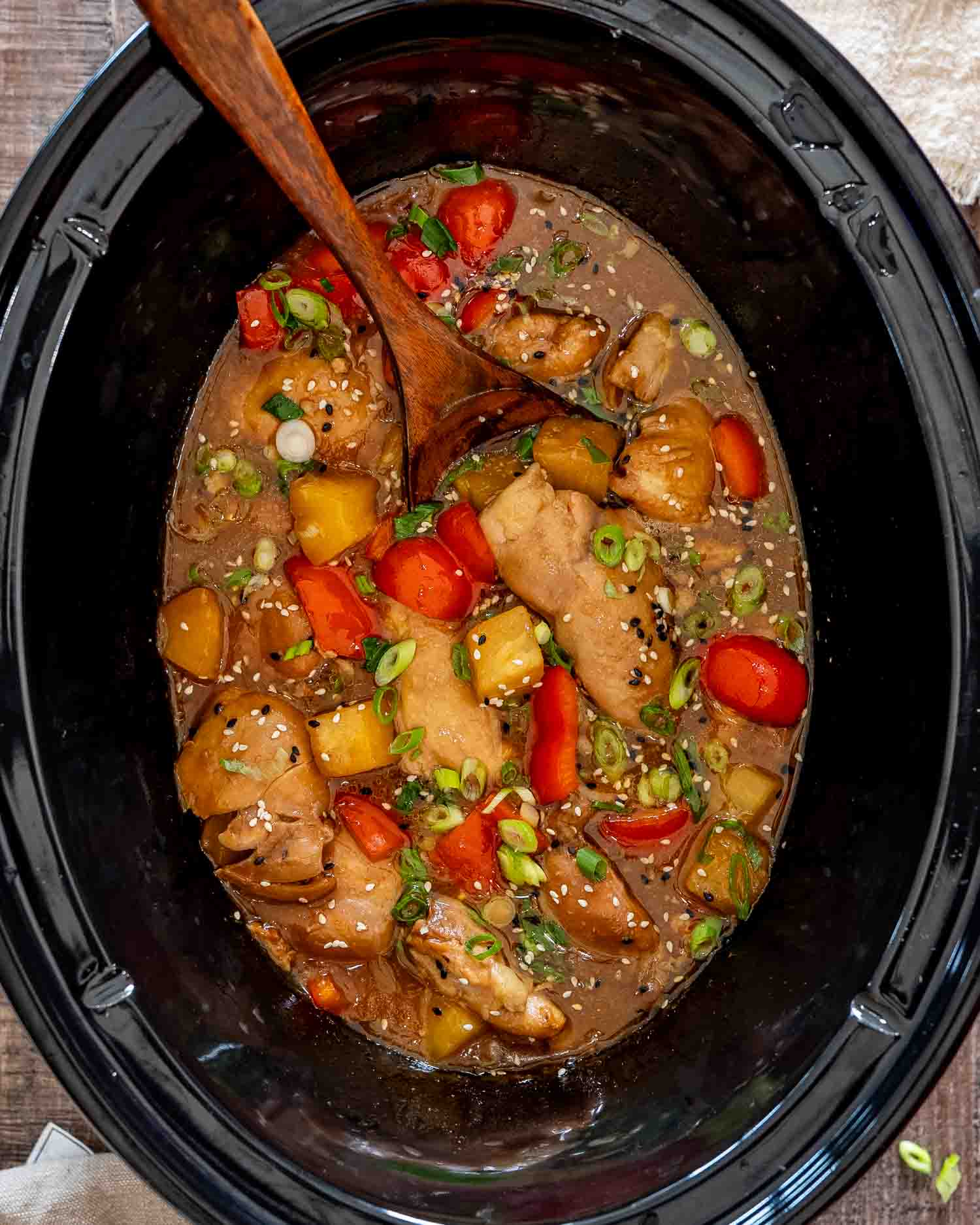 Slow cooker Hawaiian chicken with pineapple and red peppers simmers in a black crockpot beside a wooden serving spoon.