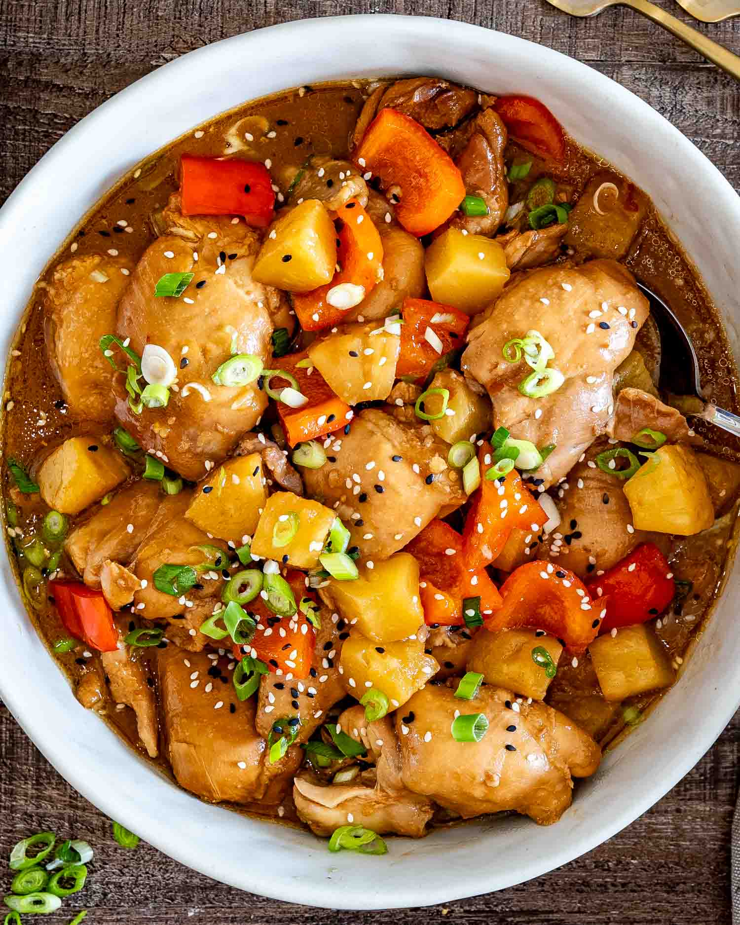 Overhead view of Hawaiian chicken thighs with pineapple, red peppers, green onions, and sesame seeds in a white bowl.
