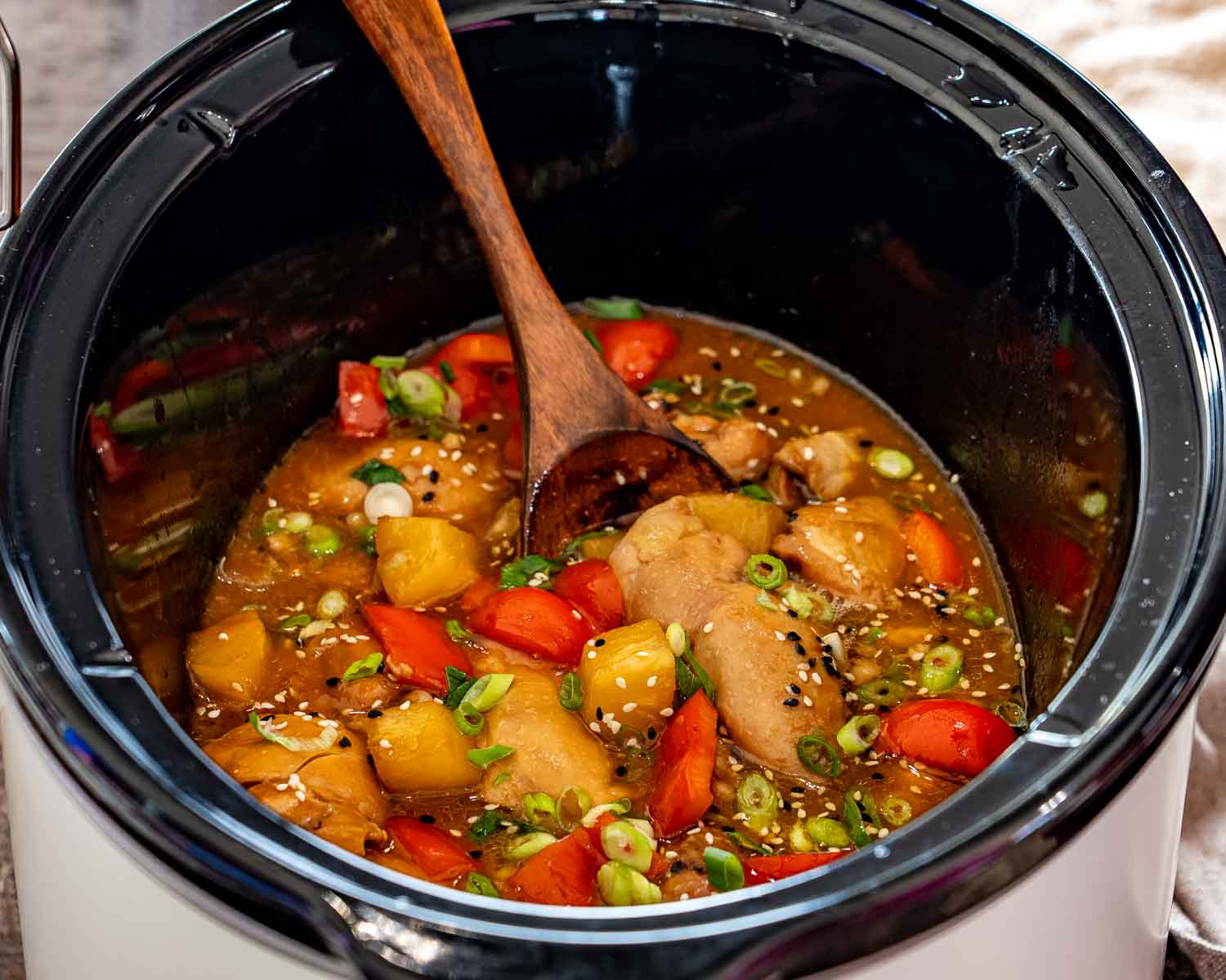 Finished Hawaiian chicken with pineapple and red peppers sits inside a slow cooker with a wooden serving spoon.