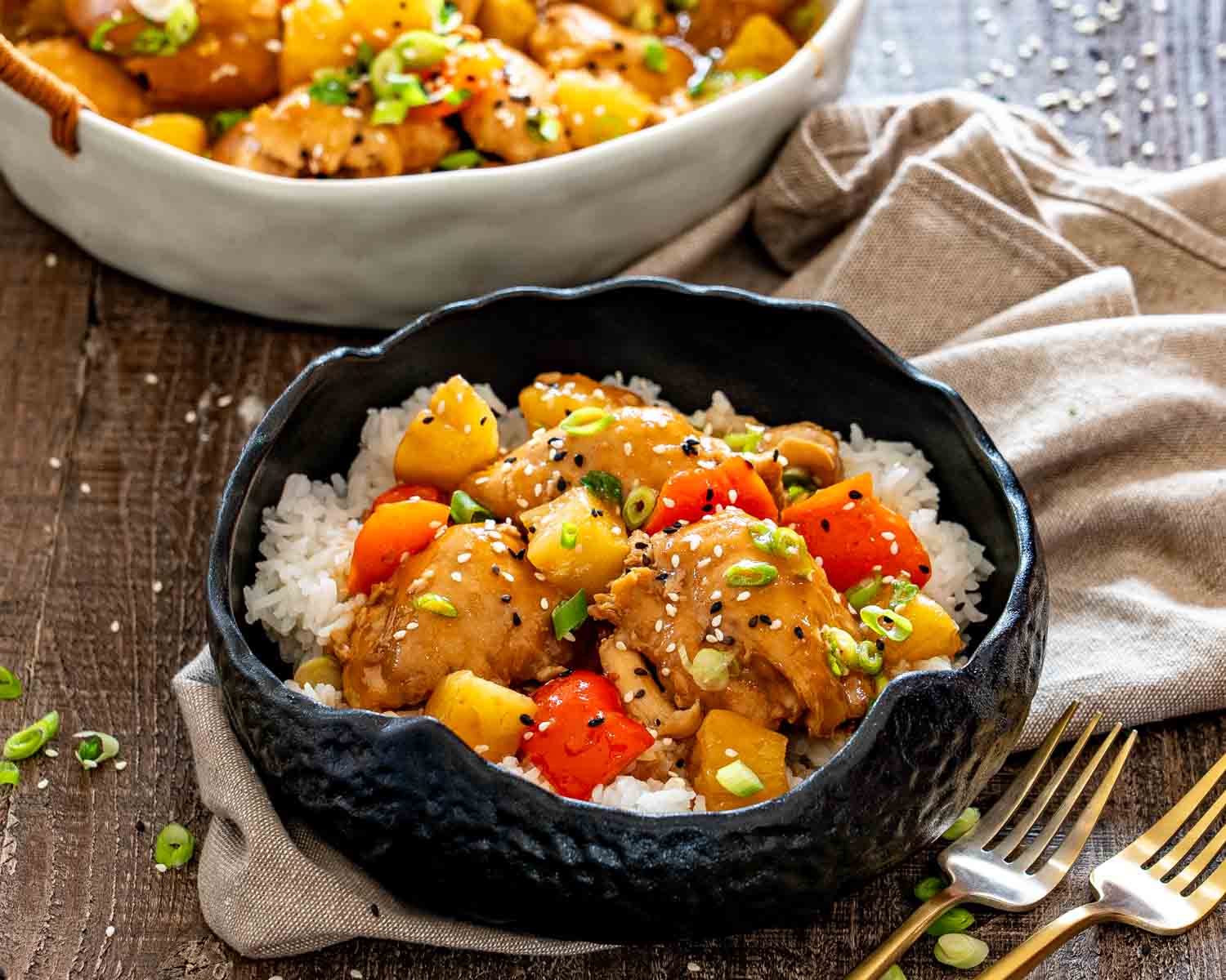 Hawaiian chicken with pineapple and red peppers served over rice in a black bowl beside a white serving dish.