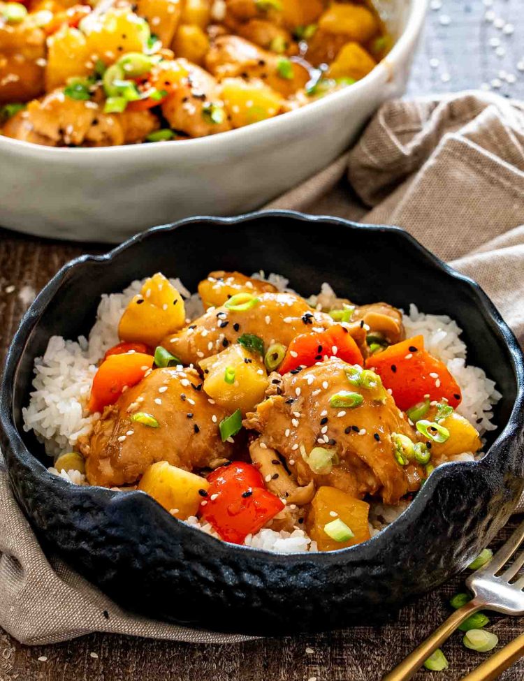 Close-up of glazed Hawaiian chicken, pineapple, and red peppers served over rice and garnished with green onions and sesame seeds.