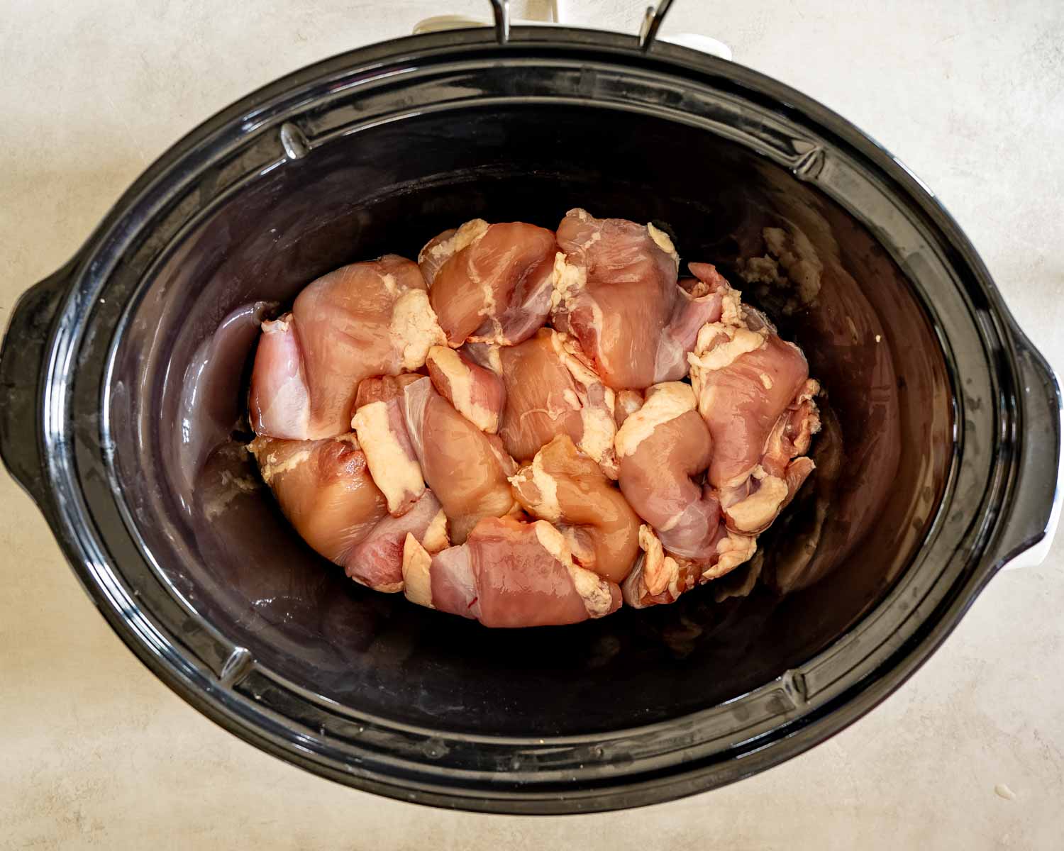 Raw boneless chicken thighs arranged inside a black slow cooker before adding the Hawaiian sauce and remaining ingredients.