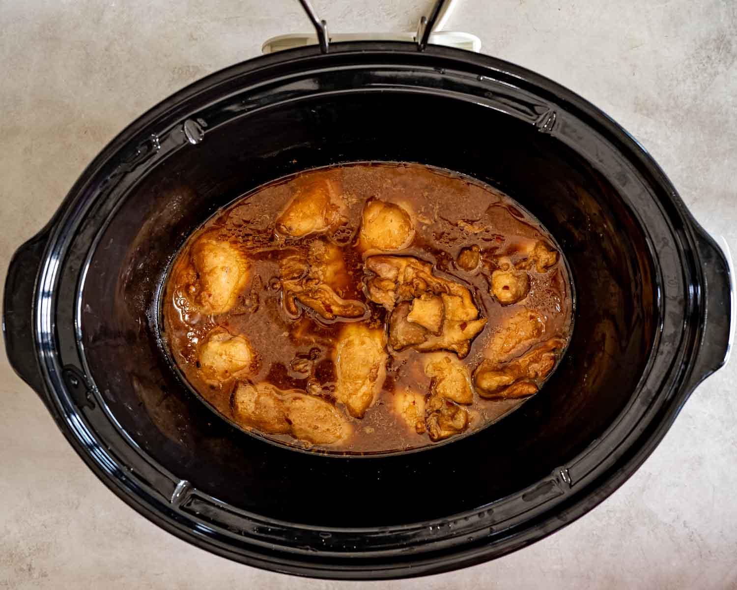 Partially cooked chicken thighs simmer in a rich brown Hawaiian sauce inside a black slow cooker.