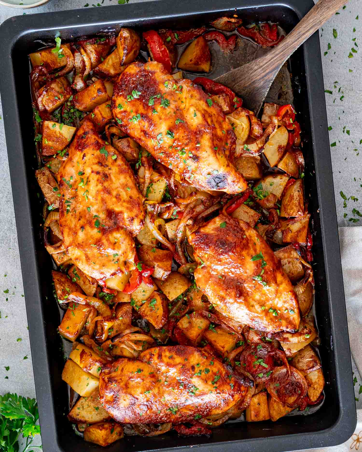 Honey garlic chicken breast lifted with a wooden spatula above roasted potatoes, peppers, and onions in a baking pan.