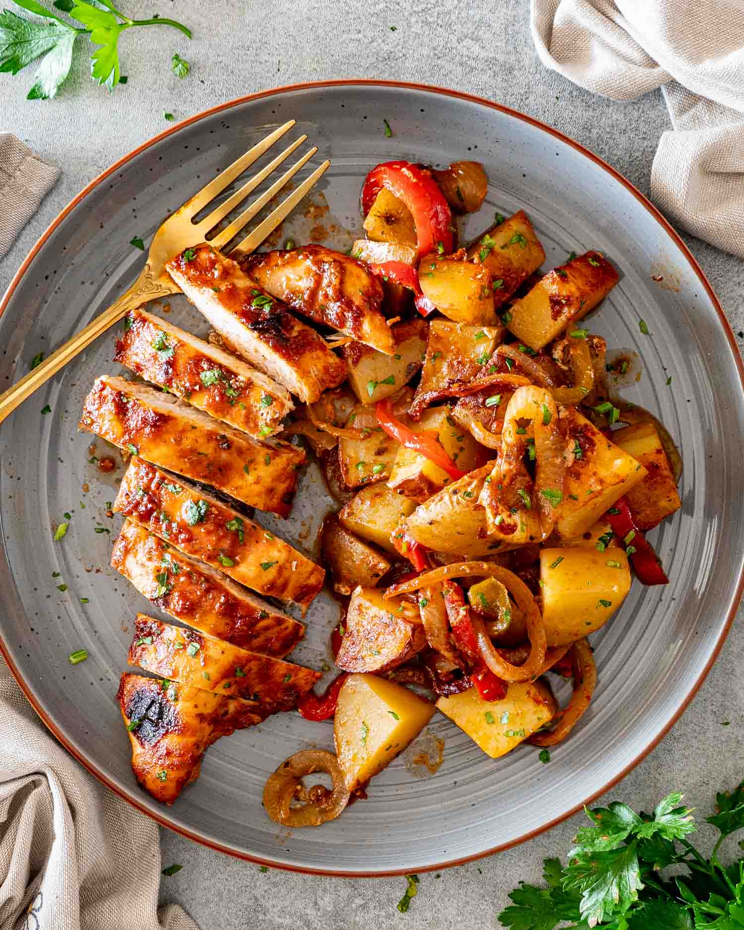 Overhead view of sliced honey garlic chicken beside roasted potatoes, peppers, and onions on a parsley garnished gray plate.
