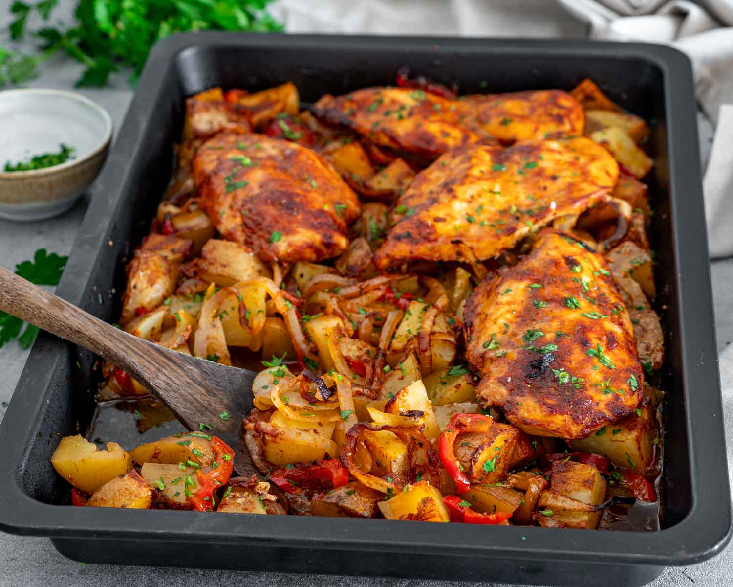 Angled view of honey garlic chicken and roasted vegetables in a black baking pan, with a wooden serving spoon.