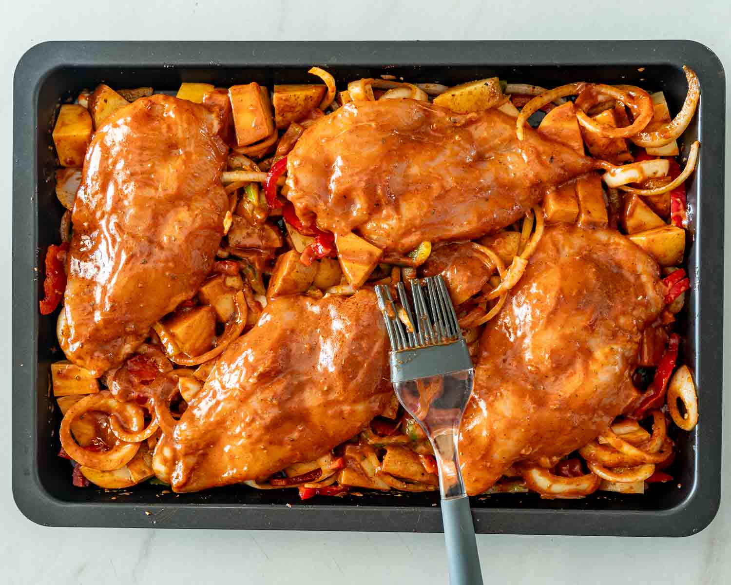Marinated chicken breasts arranged over seasoned potatoes, peppers, and onions in a baking pan with a basting brush.