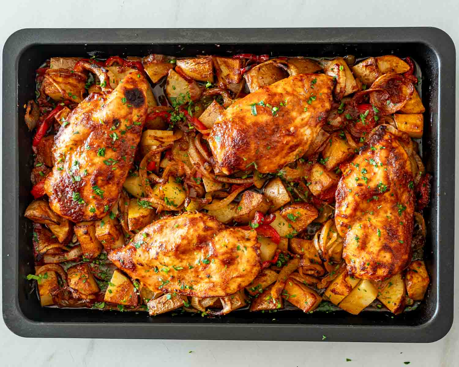 Overhead view of four golden honey garlic chicken breasts baked over potatoes, peppers, and onions, garnished with parsley.